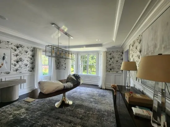 A wide-angle view of a dining room showing the elegant wallpaper installation with a black and white magnolia pattern, wainscoting, a gold geometric chandelier, and a large central oval table.